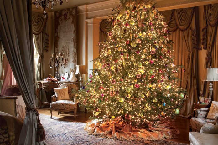An image of grandeur in an open bedroom adorned with antiques, rugs, and tapestries, this tree is dotted with hundreds of glowing orbs in shades of gold, cherry red, and Granny Smith green.