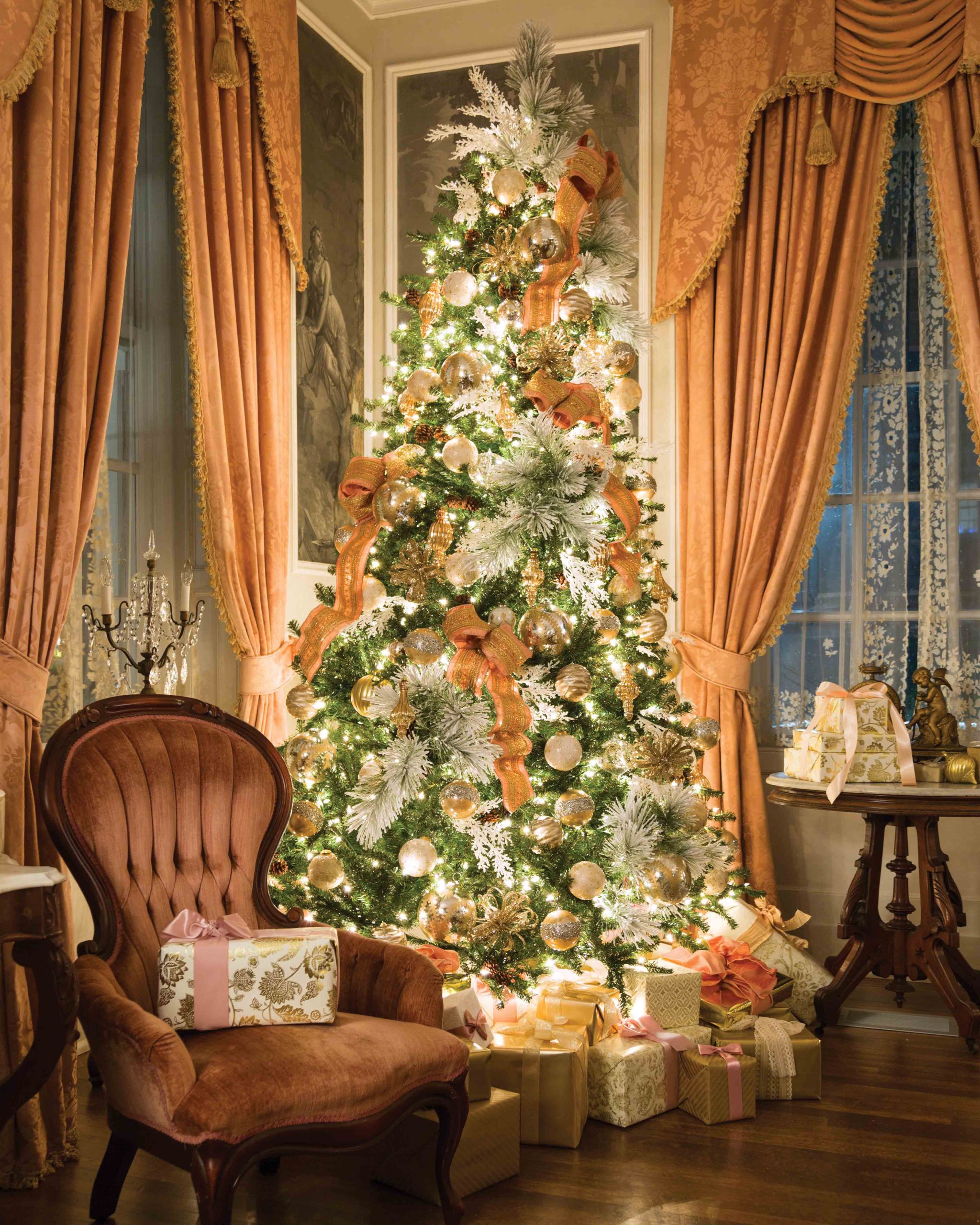 Against a backdrop of coral-hued curtains and artistic panels, an evergreen with frosted branches is adorned with pink and gold ribbons, white, silver, and gold ornaments, and shimmering lights.