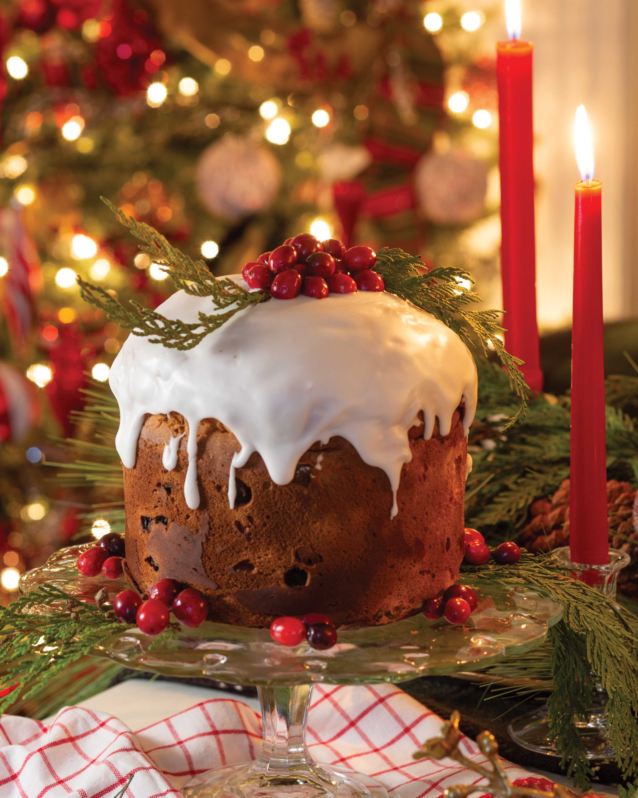 Against a backdrop of glowing lights on the tree, a glazed panettone cake dressed with cranberries shines in the light of two red candlesticks