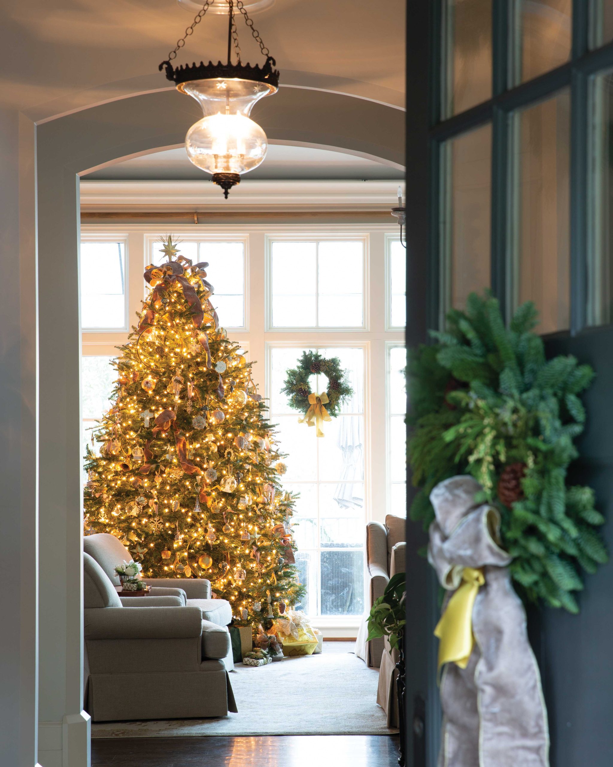 Through the front door, dressed with a beribboned wreath, the living room and its grand gold Christmas tree can be seen standing proud before a wall of windows.