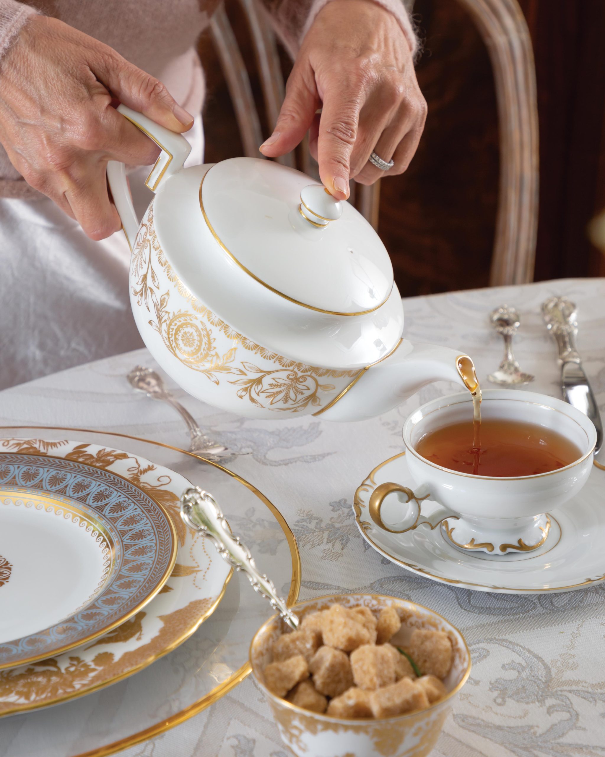A pair of hands carefully pours from a gold-and-white teacup at a table set for afternoon tea.