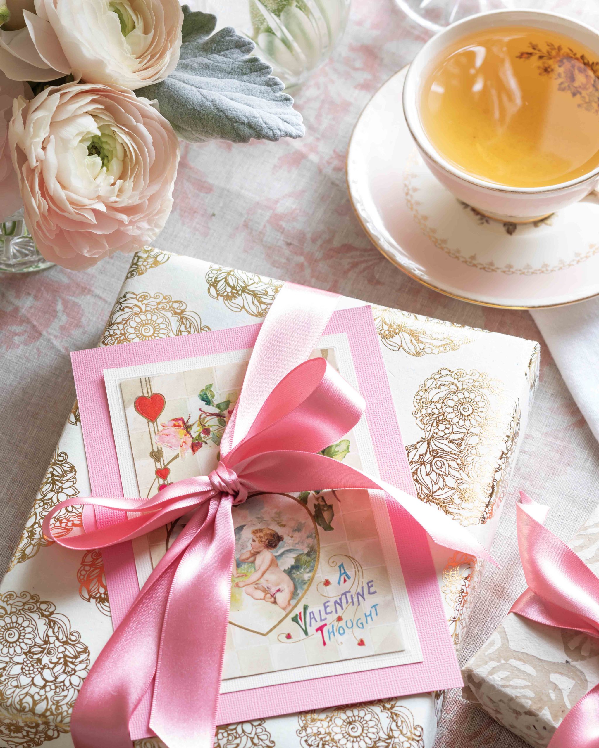 A gift tied with a bright pink ribbon and set beside a rose-colored teacup conceals a romantic card from one’s Valentine tucked into the packaging.