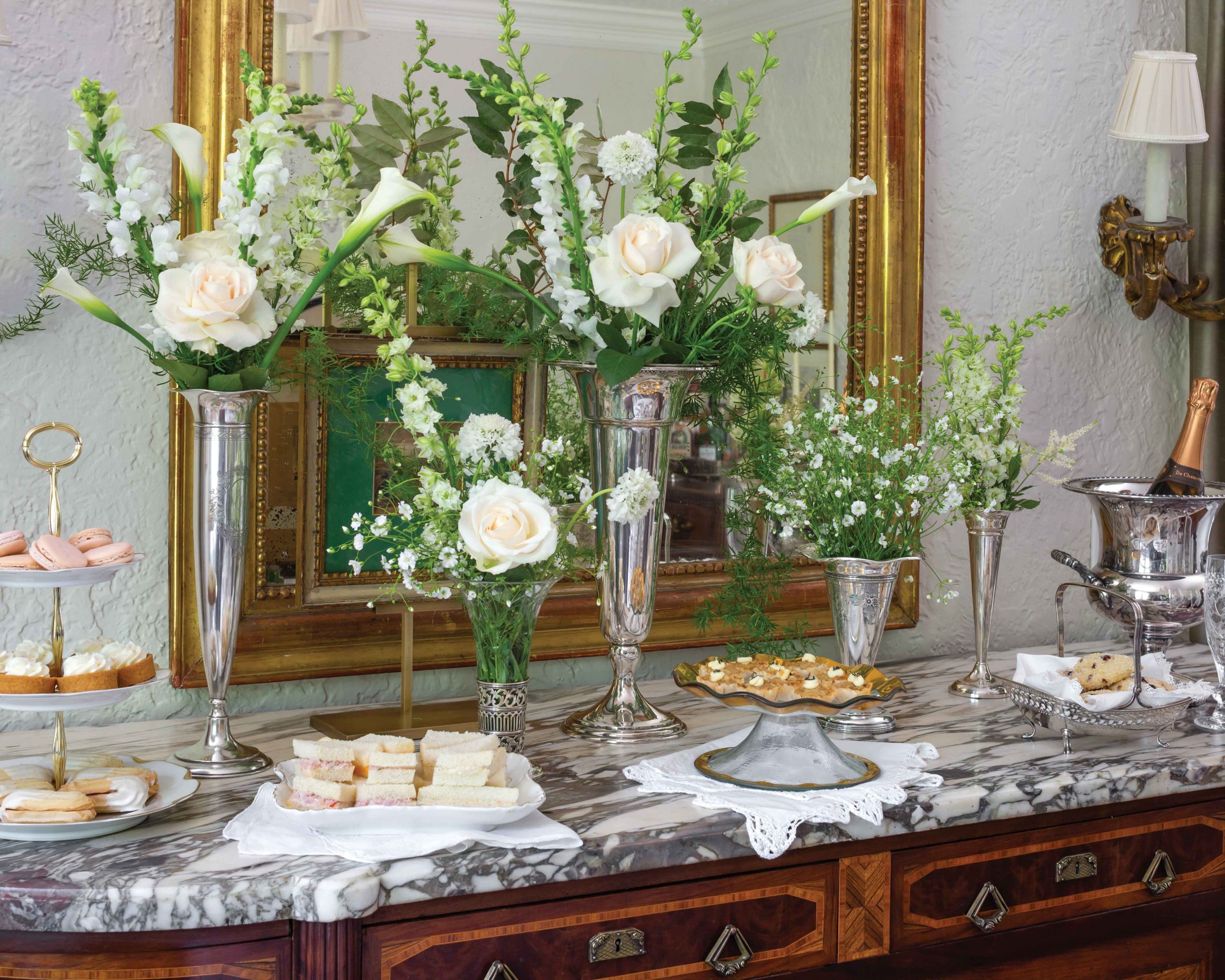 A buffet is set with trays of small dishes perfect for teatime, with a background of white roses displayed in silver vases.