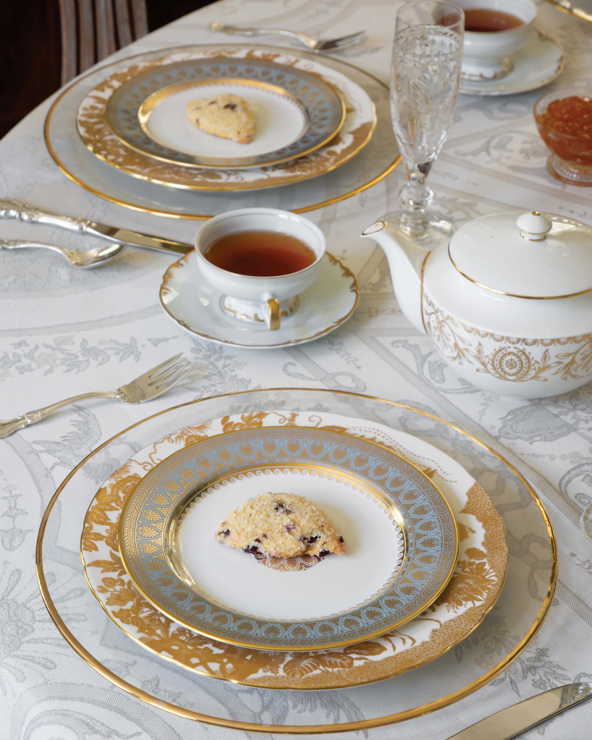 A table set for afternoon tea features gold-and-white china and a sweet scone ready for tasting.