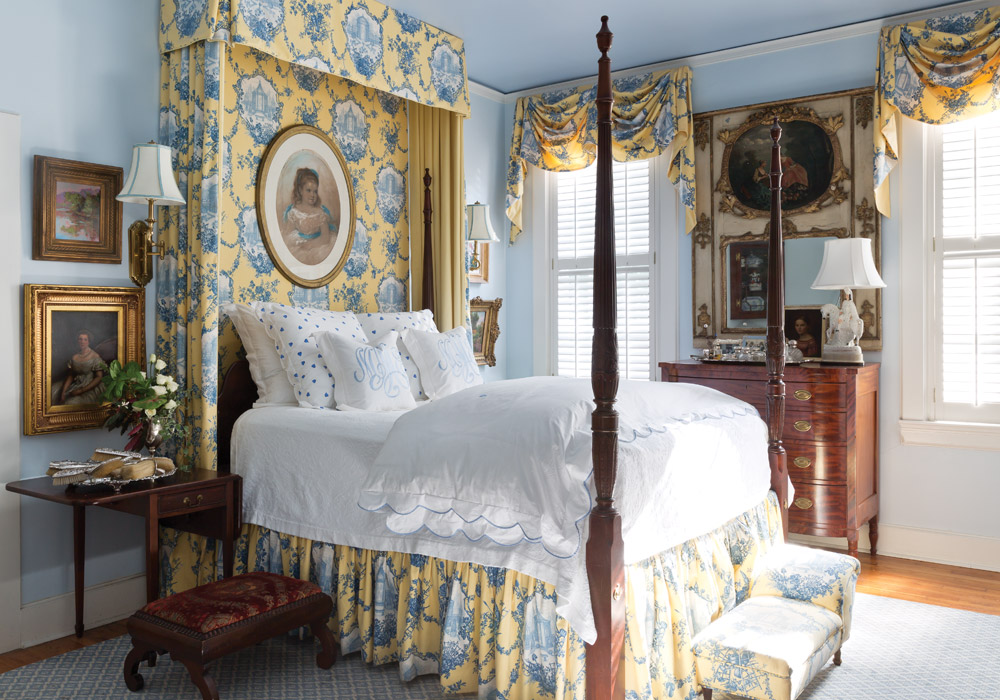A blue bedroom features a four-poster bed dressed in yellow printed fabrics and bright white linens.