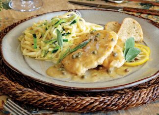 Served with a glass of white wine, zucchini noodles and chicken piccata are arranged on a plate beside slices of crunchy ciabatta.