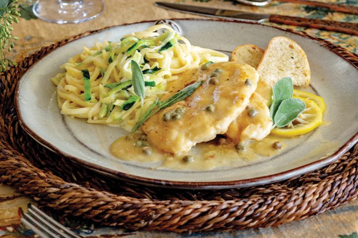 Served with a glass of white wine, zucchini noodles and chicken piccata are arranged on a plate beside slices of crunchy ciabatta.