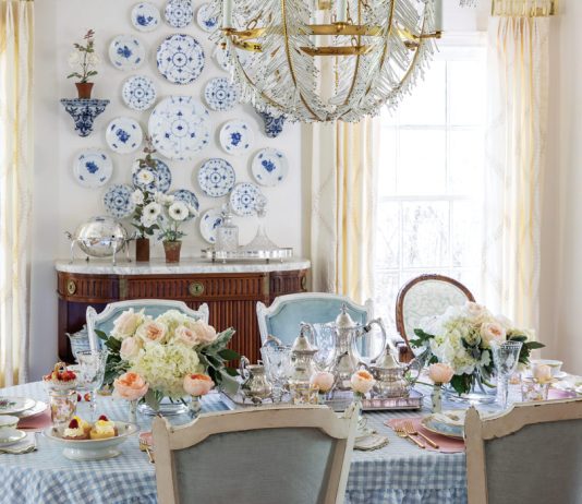 A rectangular dining table in Nicola Bathie McLaughlin’s Texas home displays a blue-and-white checked tablecloth, colorful china, and bouquets of pastel flowers.
