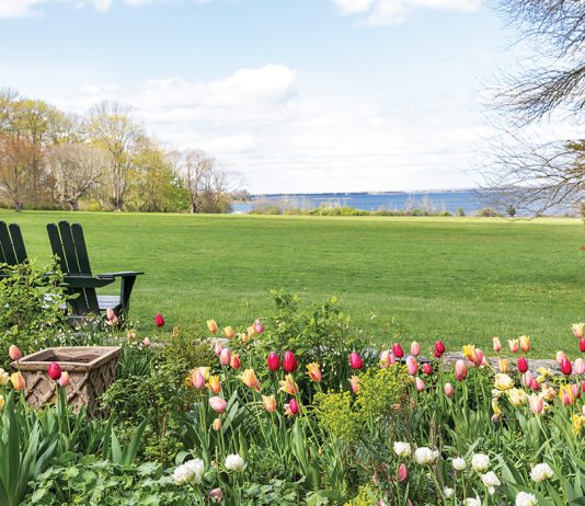 Two slender Adirondack chairs sit at the edge of a tulip garden and a verdant field, looking out onto the bay.