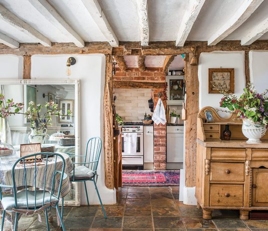 From Coast to Countryside: A breakfast nook with blue chairs sits beneath whitewashed wood ceilings and storied wooden furniture, with a peek through the open doorway into the cozy kitchen.